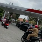 Pengendara sepeda motor antre di SPBU beratap merah putih, satu skuter melintas di halaman berpaving