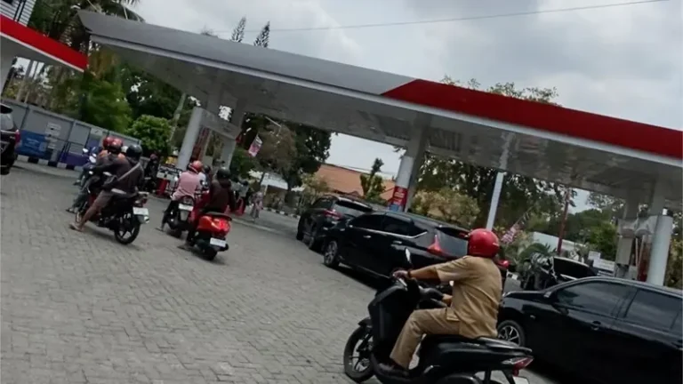 Pengendara sepeda motor antre di SPBU beratap merah putih, satu skuter melintas di halaman berpaving